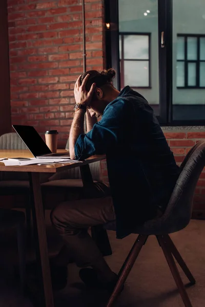 Frustrado empresario tocando la cabeza y sentado cerca de la computadora portátil con pantalla en blanco - foto de stock