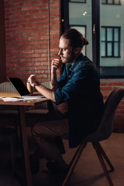 Hombre de negocios mirando reloj y sentado cerca de la computadora portátil con pantalla en blanco - foto de stock