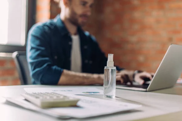 Enfoque selectivo de la botella con desinfectante de manos cerca de la mujer de negocios utilizando el ordenador portátil en la oficina - foto de stock