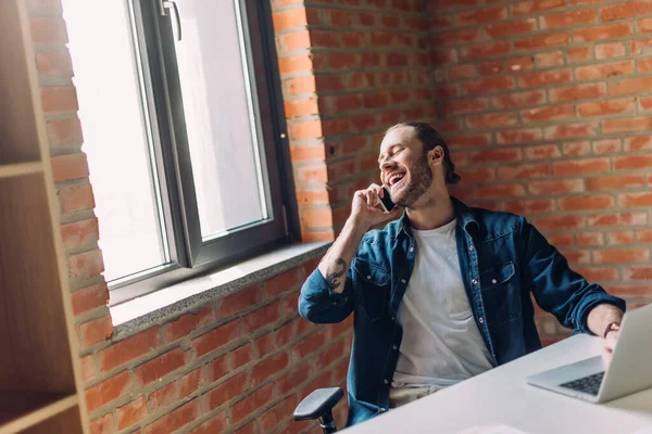 Избирательный фокус счастливого бизнесмена, разговаривающего на смартфоне и использующего ноутбук в офисе — стоковое фото