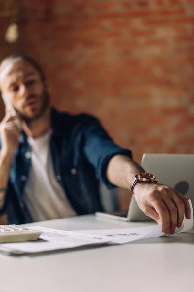 Selective focus of businessman talking on smartphone and taking paper near laptop in office — Stock Photo