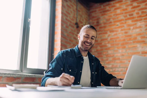 Foco seletivo de homem de negócios feliz escrevendo no caderno perto do computador portátil no escritório — Fotografia de Stock