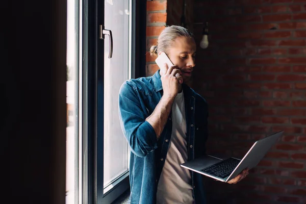 Selektiver Fokus des Geschäftsmannes, der auf Laptop schaut und am Fenster mit dem Smartphone spricht — Stockfoto
