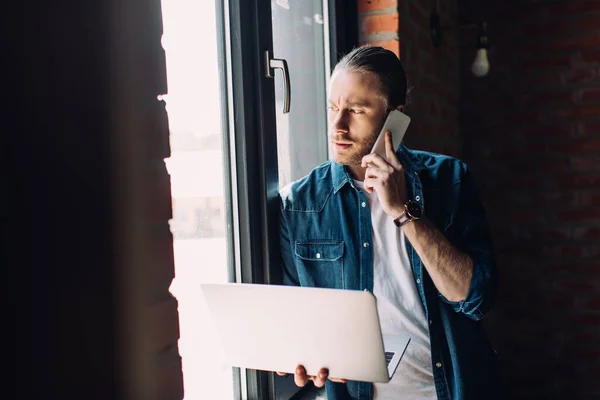 Selektiver Fokus des bärtigen Geschäftsmannes, der Laptop in der Hand hält und am Fenster mit dem Smartphone spricht — Stockfoto
