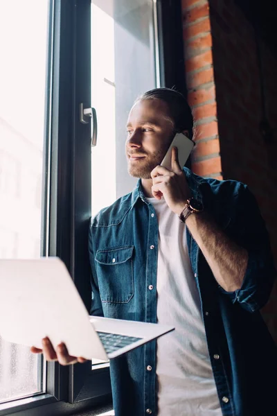 Enfoque selectivo de hombre de negocios sosteniendo el ordenador portátil y hablando en el teléfono inteligente cerca de la ventana - foto de stock