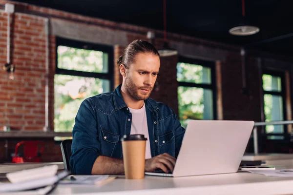 Избирательный фокус бизнесмена, смотрящего на ноутбук рядом с бумажной чашкой на столе — стоковое фото