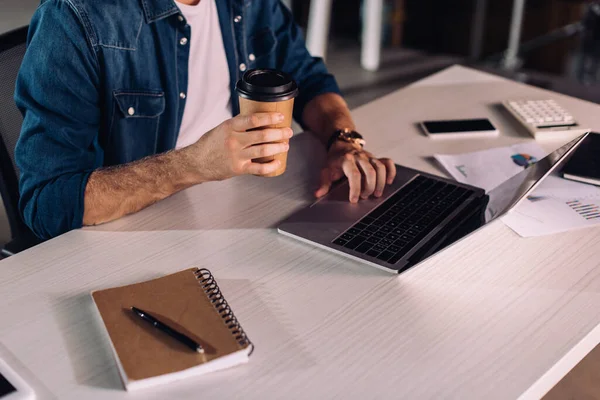 Обрезанный вид бизнесмена, держащего бумажную чашку и использующего ноутбук рядом со смартфоном на столе — стоковое фото