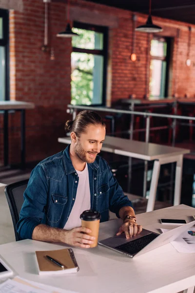 Homem de negócios feliz segurando copo de papel e usando laptop perto de smartphone na mesa — Fotografia de Stock