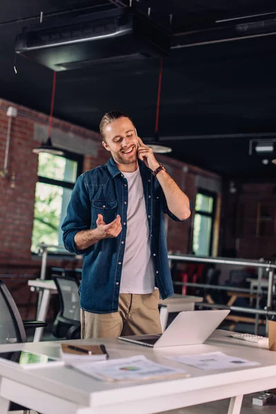 Selektiver Fokus der gut gelaunten Geschäftsfrau, die im Büro auf dem Smartphone neben dem Laptop spricht — Stockfoto