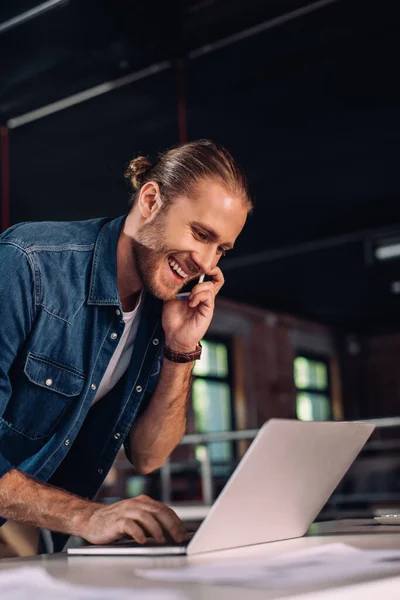 Cheerful businesswoman using laptop while talking on smartphone — Stock Photo