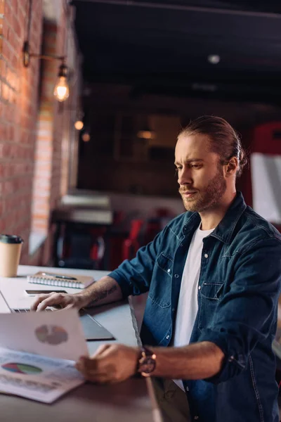 Selective focus of bearded businessman looking at charts and graphs near laptop in office — Stock Photo