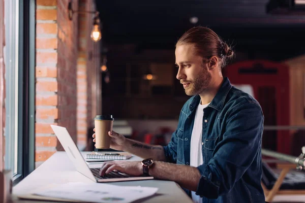 Вибірковий фокус бородатого бізнесмена, який дивиться на ноутбук і тримає одноразову чашку в офісі — стокове фото