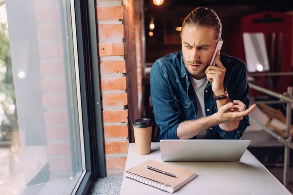 Homem de negócios falando no smartphone perto de copo de papel e laptop — Fotografia de Stock