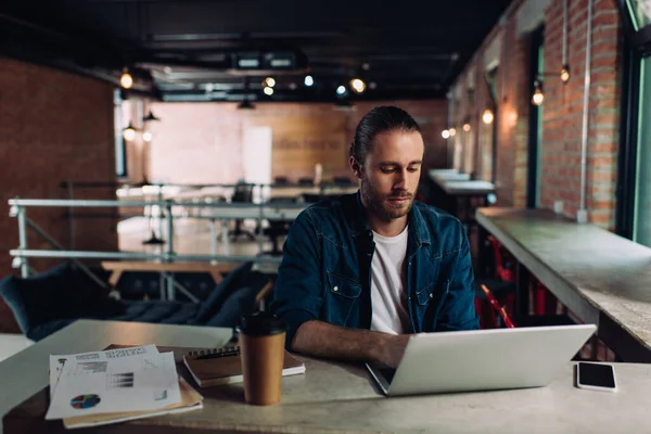 Hombre de negocios utilizando el ordenador portátil cerca de taza de papel y teléfono inteligente con pantalla en blanco en el escritorio - foto de stock