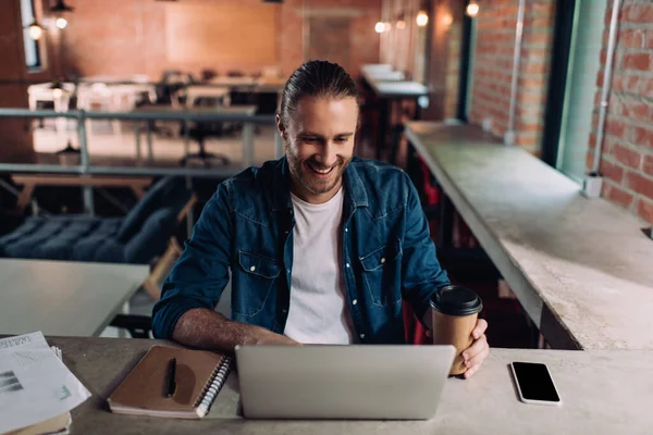Alegre hombre de negocios mirando a la computadora portátil y sosteniendo la taza de papel cerca del teléfono inteligente con pantalla en blanco - foto de stock
