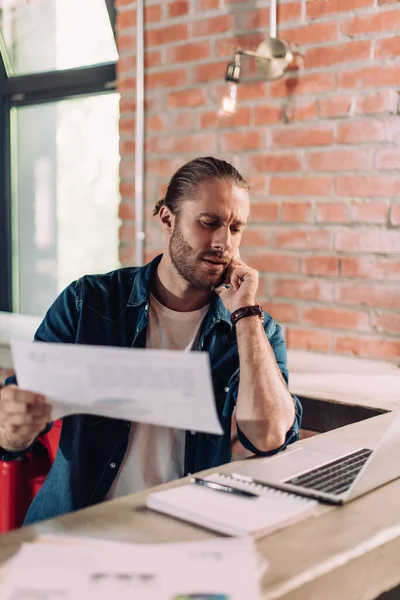 Selektiver Fokus des Geschäftsmannes, der auf den Laptop schaut, während er Diagramme und Grafiken hält und auf dem Smartphone spricht — Stockfoto