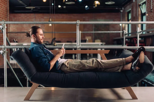 Vue latérale de l'homme d'affaires se refroidissant sur le canapé et utilisant un smartphone dans le bureau — Photo de stock
