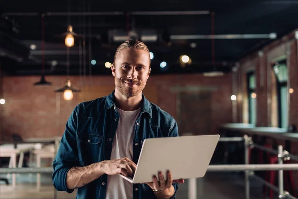 Sonriente hombre de negocios sosteniendo y utilizando el ordenador portátil en la oficina moderna - foto de stock
