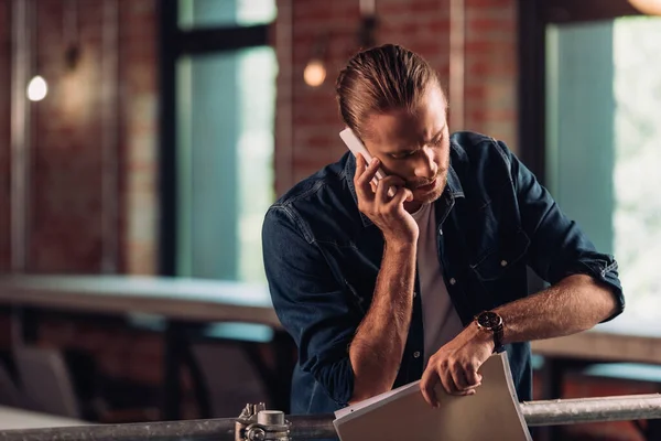 Bärtiger Geschäftsmann spricht auf Smartphone und schaut auf Uhr, während er Ordner im Büro hält — Stockfoto