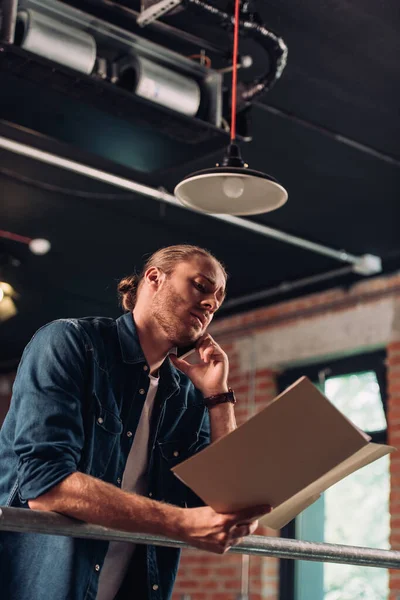 Low angle view of handsome businessman talking on smartphone and holding folder in office — Stock Photo