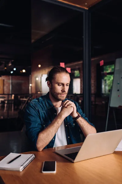 Bearded businessman with clenched hands looking at laptop near notebook and smartphone with blank screen — Stock Photo