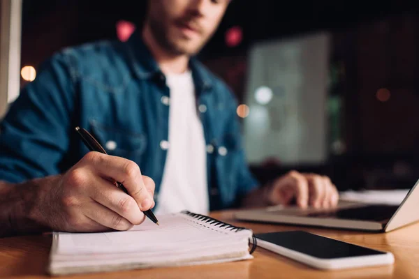 Vista ritagliata di uomo d'affari scrittura in notebook vicino smartphone e laptop — Foto stock