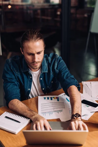 Hombre de negocios enfocado utilizando el ordenador portátil cerca de teléfono inteligente con pantalla en blanco, gráficos y gráficos en el escritorio - foto de stock