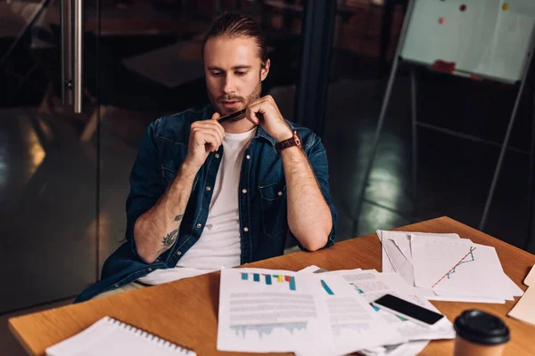 Селективный фокус бородатого бизнесмена, держащего ручку и смотрящего на графики и графики на столе — стоковое фото