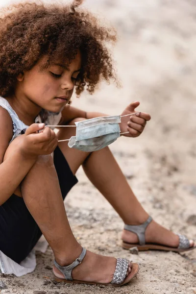 Curly african american kid holding dirty medical mask while sitting on ground — Stock Photo