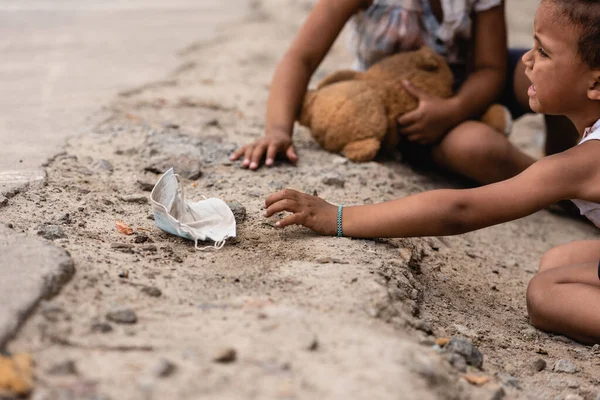 Messa a fuoco selettiva del povero ragazzo africano americano piangendo mentre raggiungeva sporca maschera medica vicino sorella a terra — Foto stock