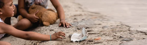Cultivo panorámico de niño afroamericano pobre llorando mientras que alcanza máscara médica sucia cerca de la hermana en el suelo - foto de stock