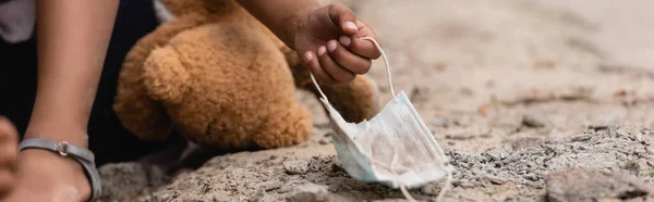 Panoramisches Konzept des armen afrikanisch-amerikanischen Kindes, das schmutzige medizinische Maske in der Nähe von Teddybär auf dem Boden berührt — Stockfoto