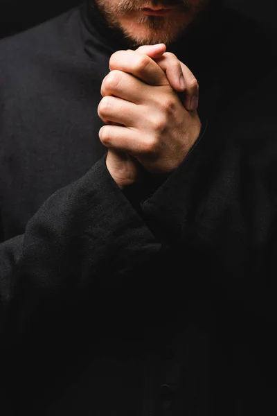 Vista recortada del sacerdote con las manos apretadas rezando aislado en negro - foto de stock