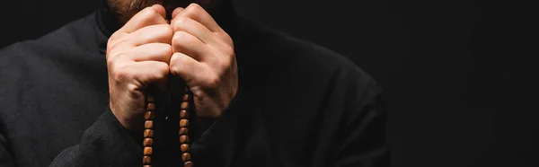 Cultivo panorámico de sacerdote sosteniendo cuentas de rosario en manos aisladas en negro - foto de stock