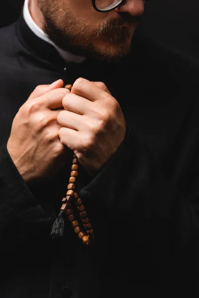 Visão cortada de padre barbudo segurando contas de rosário em mãos e orando isolado em preto — Fotografia de Stock