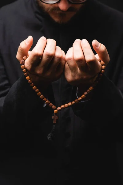 Vista recortada del pastor sosteniendo cuentas de rosario en las manos y rezando aislado en negro - foto de stock