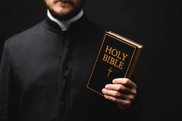 Vista recortada de sacerdote sosteniendo la Biblia santa aislado en negro - foto de stock