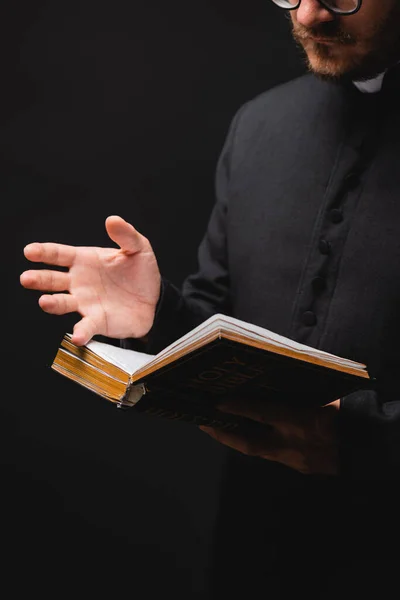 Vista cortada de padre segurando bíblia sagrada e gesto isolado em preto — Fotografia de Stock