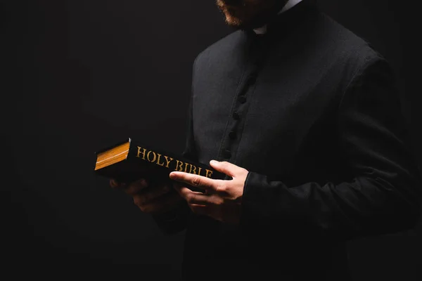 Vue recadrée du prêtre barbu tenant la bible sainte dans les mains isolées sur noir — Photo de stock