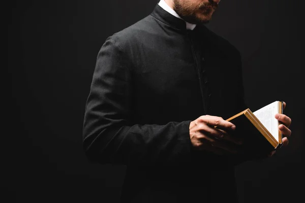 Partial view of bearded priest holding holy bible in hands isolated on black — Stock Photo