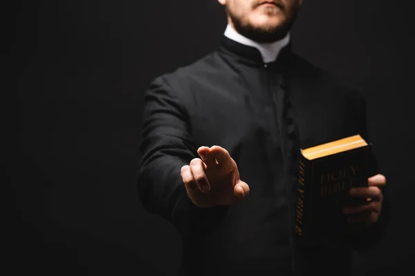 Foco seletivo do padre segurando bíblia sagrada enquanto gesticulando isolado no preto — Fotografia de Stock