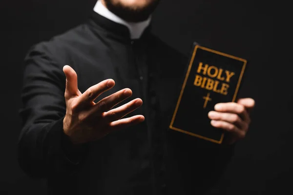 Enfoque selectivo del pastor sosteniendo la Biblia sagrada mientras gesticulaba aislado en negro - foto de stock