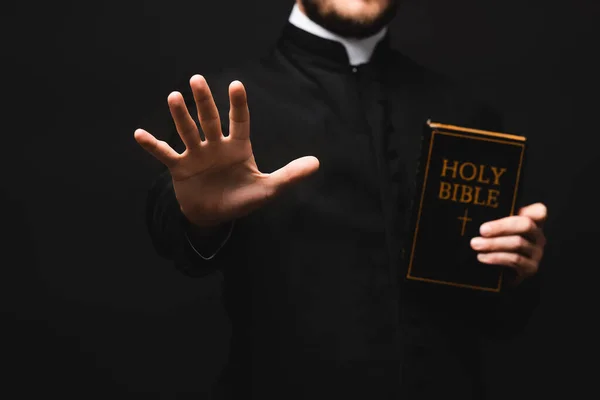 Vista cortada de padre segurando bíblia sagrada enquanto gesticulando isolado em preto — Fotografia de Stock