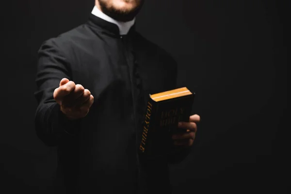 Cropped view of priest holding holy bible while showing fist isolated on black — Stock Photo