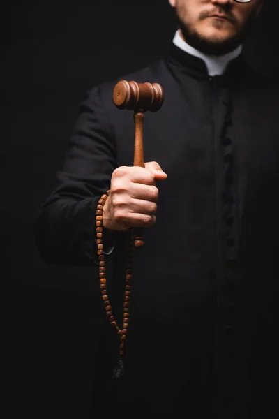 Cropped view of priest holding wooden gavel and rosary beads isolated on black — Stock Photo