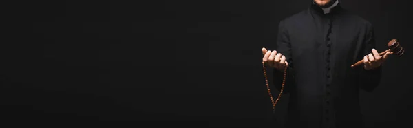 Panoramic shot of priest holding wooden gavel and rosary beads in hands isolated on black — Stock Photo