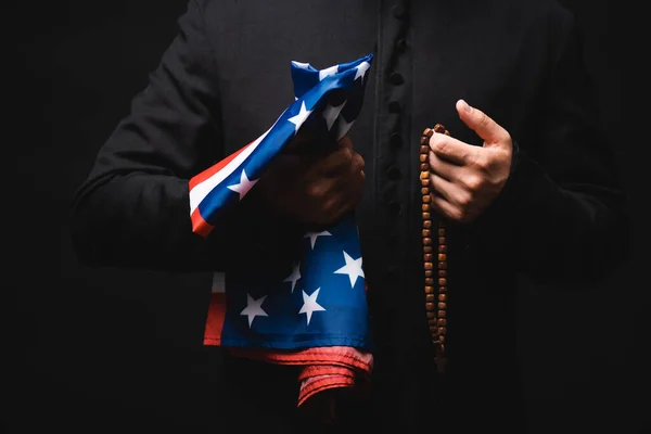 Abgeschnittene Ansicht des Priesters mit amerikanischer Flagge und Rosenkranzperlen in den Händen isoliert auf schwarz — Stockfoto