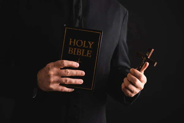 Abgeschnittene Ansicht eines Priesters mit Kreuz und heiliger Bibel isoliert auf schwarz — Stockfoto