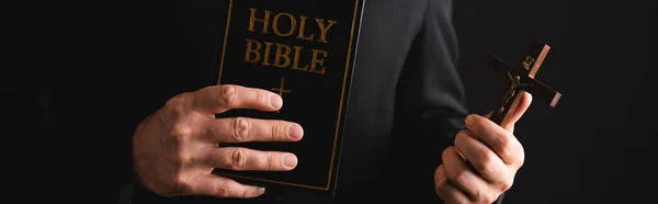 Panoramic concept of priest holding cross and holy bible isolated on black — Stock Photo