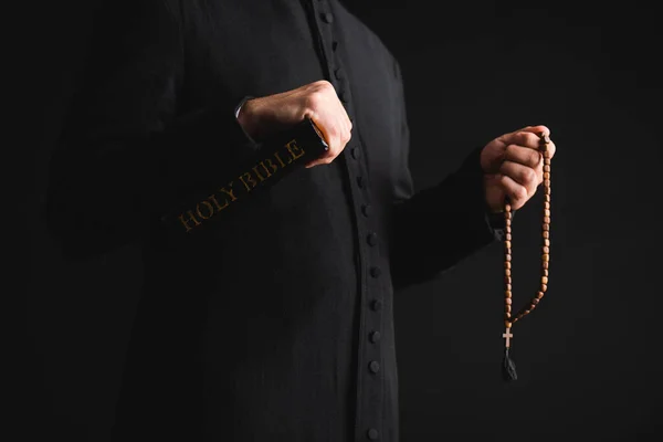 Vista recortada del pastor sosteniendo la sagrada Biblia y rosarios en las manos aisladas en negro - foto de stock